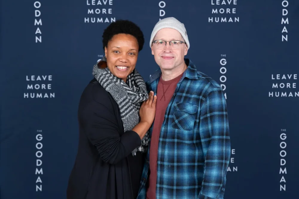 A woman and a man standing together in front of a blue backdrop featuring the repeated text “Leave More Human” and “The Goodman.”