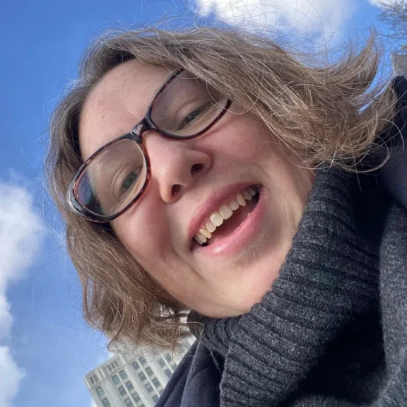 A person wearing glasses and a dark turtleneck sweater smiling at the camera outdoors, with a building and blue sky in the background.