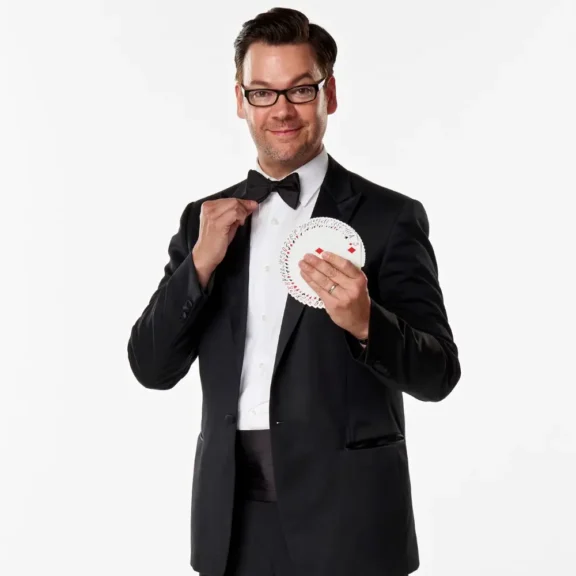 Man in a black tuxedo and glasses holds a fanned deck of playing cards in one hand and adjusts his bow tie with the other, standing against a plain white background.