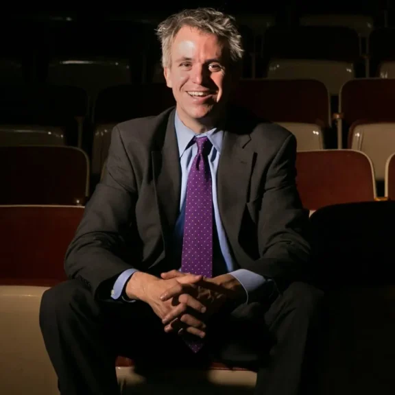A man in a suit and purple tie sits smiling with hands clasped, in a dimly lit, empty auditorium with brown and beige seats.