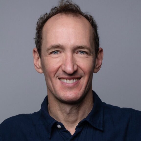 A middle-aged man with short, thinning brown hair smiles at the camera against a plain gray background. He is wearing a dark blue shirt.
