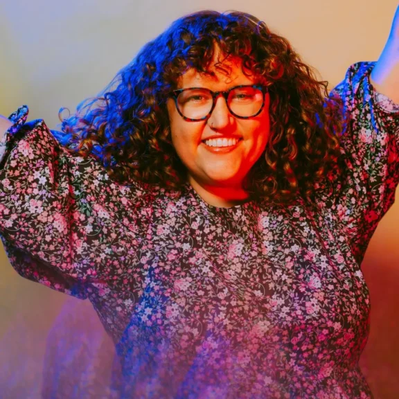 A person with curly hair and glasses smiles with arms raised, wearing a floral blouse against a colorful, softly lit background.