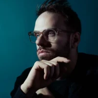A man with short hair, a beard, and clear-frame glasses looks upward pensively with his chin resting on his hand against a dark teal background.