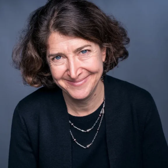 A woman with short, curly brown hair and blue eyes smiles at the camera. She is wearing a black top and a silver necklace, against a plain dark background.