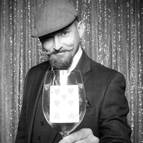 A man in a suit and cap holds a wine glass containing a playing card in front of a glittery curtain backdrop.