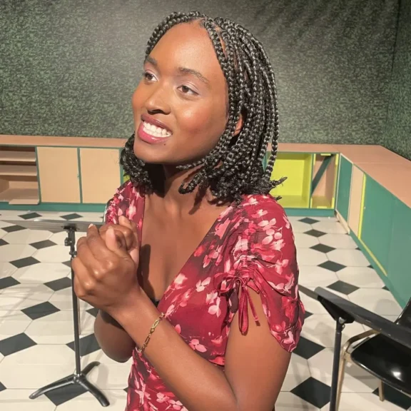 A woman with braided hair wearing a red floral dress stands indoors, smiling with her hands clasped in front of her. A microphone stand and chairs are visible in the background.