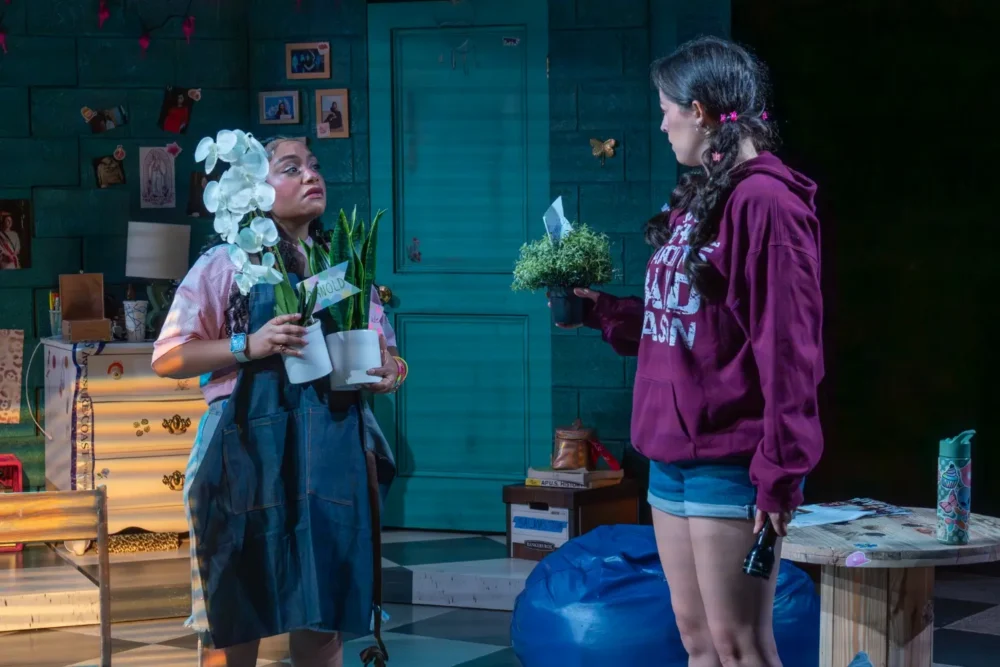 Two women stand indoors facing each other, each holding a potted plant. One wears an apron and glasses, while the other is in a maroon hoodie and shorts. The room is decorated with various items.