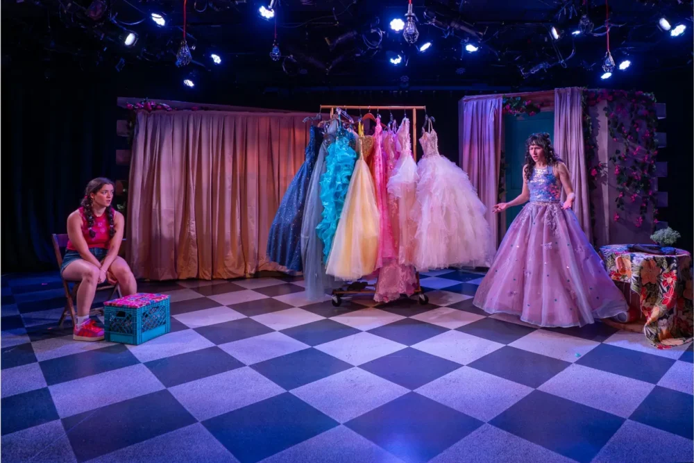 Two women are on a checkered stage; one sits by a box, while the other stands in a ball gown near a rack of elaborate dresses, surrounded by draped curtains and stage lighting.