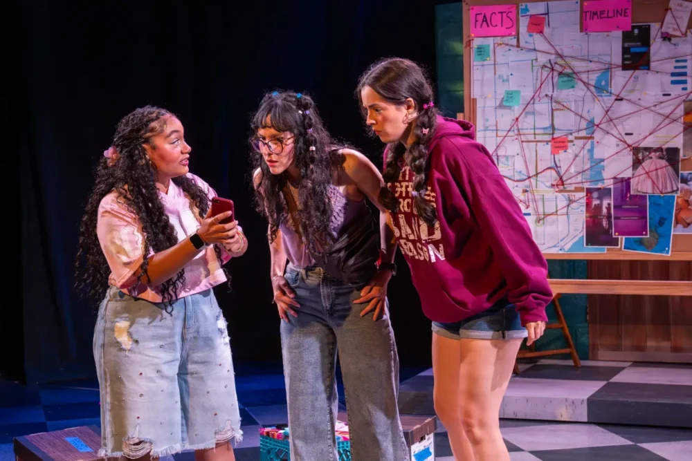 Three young women stand closely together looking at a smartphone, with a large evidence board visible behind them on stage.