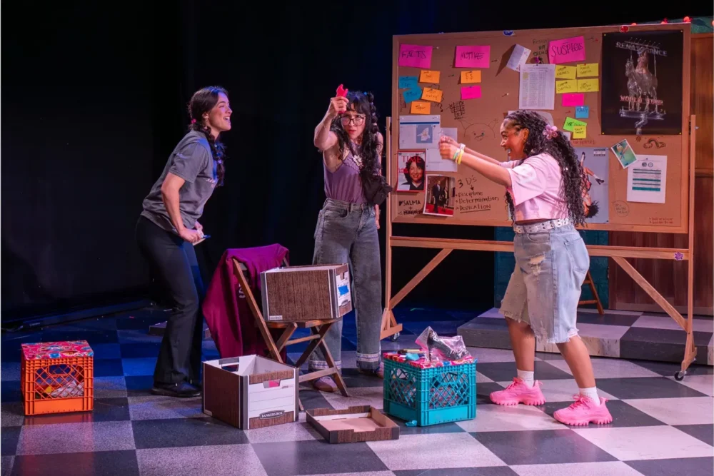 Three people interact animatedly in a room with crates and boxes, in front of a bulletin board covered with colorful notes and posters. Two people gesture with pink toy guns.