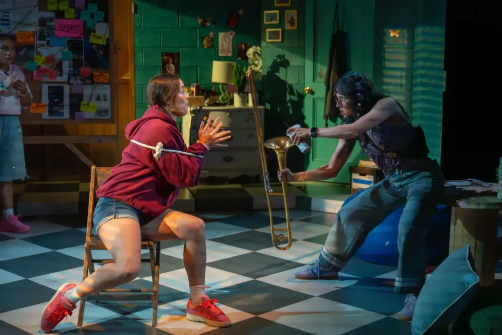 Two people face each other in a colorful room; one sits tied to a chair looking surprised, while the other points a trombone at them aggressively.