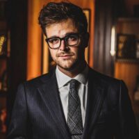 A man wearing glasses, a dark suit, and a patterned tie stands indoors, looking at the camera with a neutral expression.
