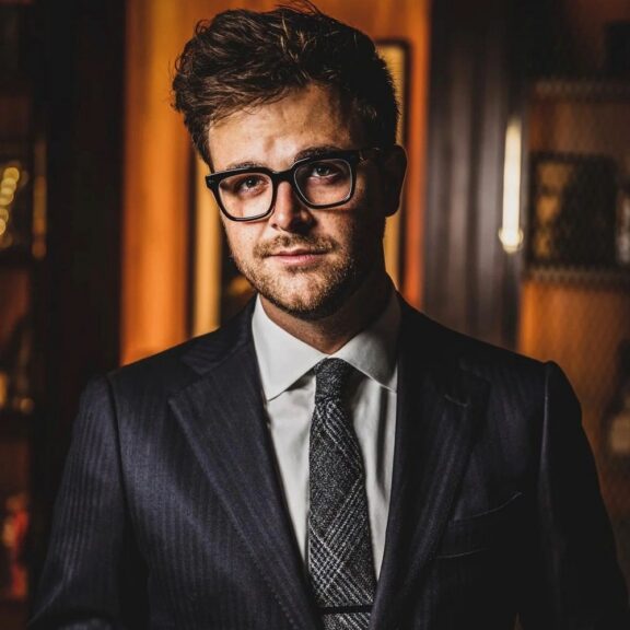 A man wearing glasses, a dark suit, and a patterned tie stands indoors, looking at the camera with a neutral expression.