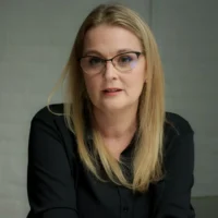A woman with long blonde hair and glasses, wearing a black shirt, sits indoors against a plain light-colored background.