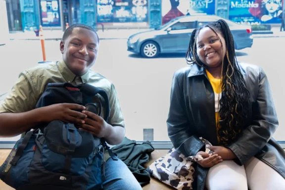 Two young people sit indoors by a large window; one holds a backpack and camera, while the other has long braids and wears a leather jacket, with a street scene visible outside.
