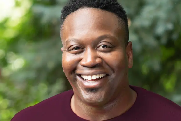 A man with short black hair smiles at the camera, wearing a maroon shirt, with greenery blurred in the background.