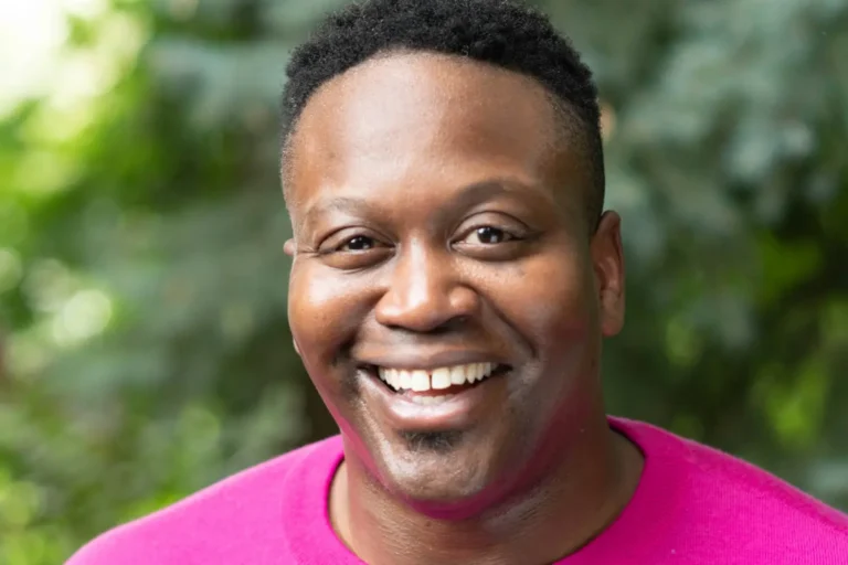 A person smiling outdoors, wearing a bright pink top, with trees and greenery in the blurred background.