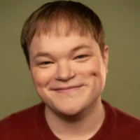 A person with short light brown hair smiles at the camera, wearing a red sweater against a plain olive green background.