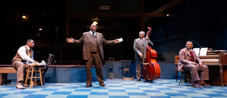 Four men in vintage suits perform on stage; one stands speaking with arms outstretched, while the others are seated or playing musical instruments, including a bass and a piano.