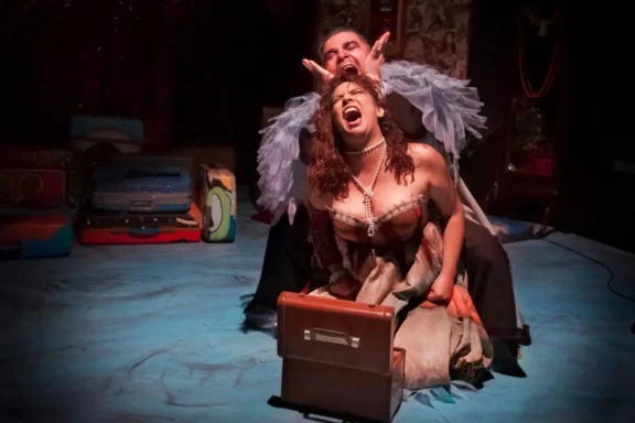 Two actors perform dramatically on stage; one sits behind the other, gesturing above her head, while she kneels and screams, clutching a brown suitcase.
