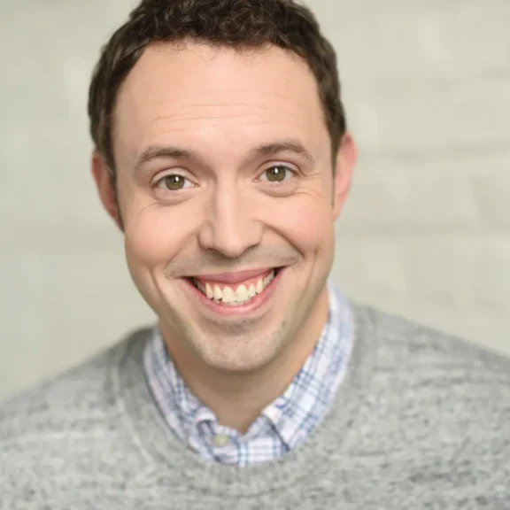 A man with short brown hair wearing a gray sweater over a checked shirt smiles at the camera against a light-colored background.