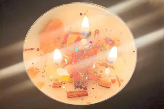 A round candle with three lit wicks, decorated with glitter, confetti, and small pieces of wood on its surface.