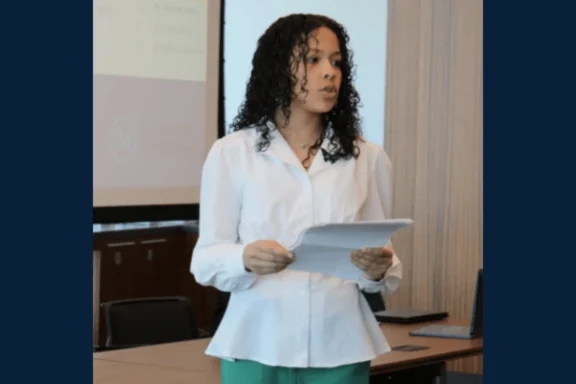 A person with curly hair, wearing a white blouse and green pants, holds papers while speaking in a conference room with a presentation screen in the background.
