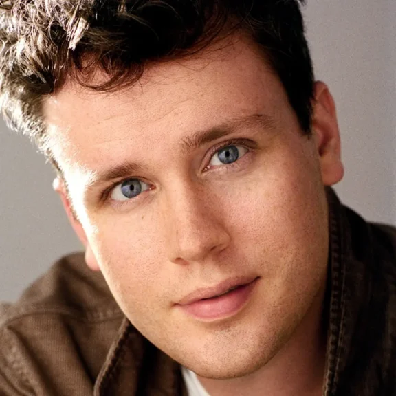 A man with short, curly brown hair and blue eyes looks directly at the camera, wearing a brown jacket and a neutral expression.