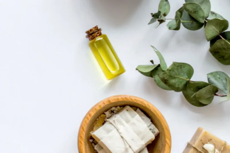 A small glass bottle of yellow oil, a wooden bowl with wrapped soaps, a bar of soap, and eucalyptus leaves arranged on a white surface.