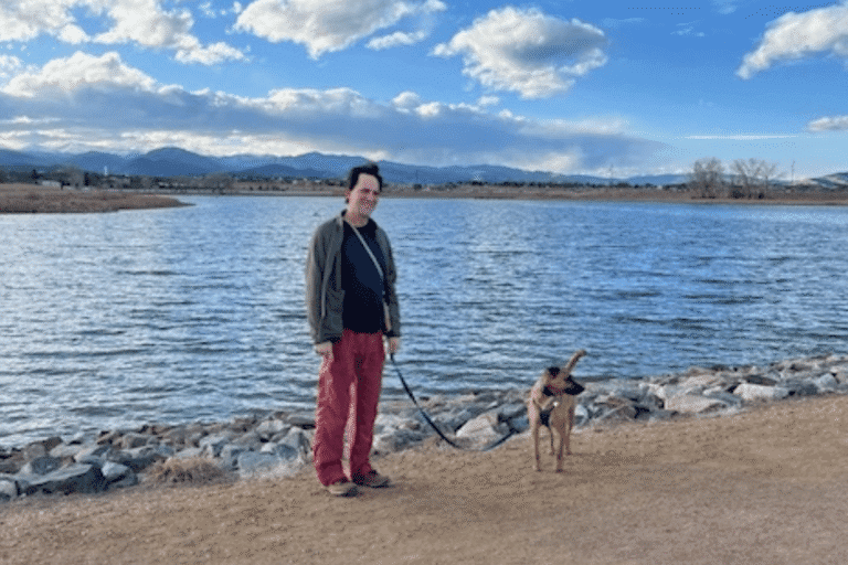 A person in red pants stands on a gravel path by a body of water, holding the leash of a brown dog. Mountains and clouds are visible in the background.