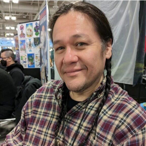A person with long dark hair in braids, wearing a plaid shirt, smiles while seated indoors at a convention or event with art displays in the background.
