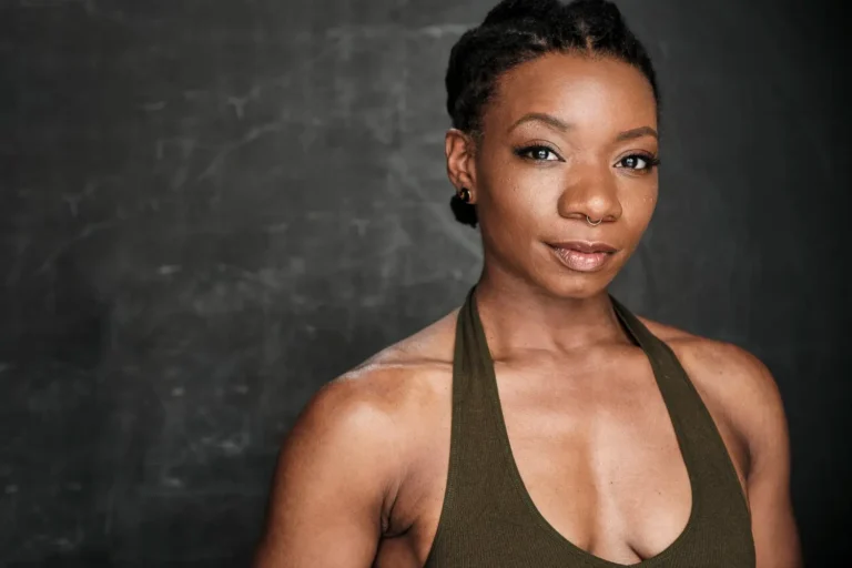 A woman with short braided hair, wearing a sleeveless olive green top, stands in front of a dark, textured background. She is looking slightly to the side with a neutral expression.