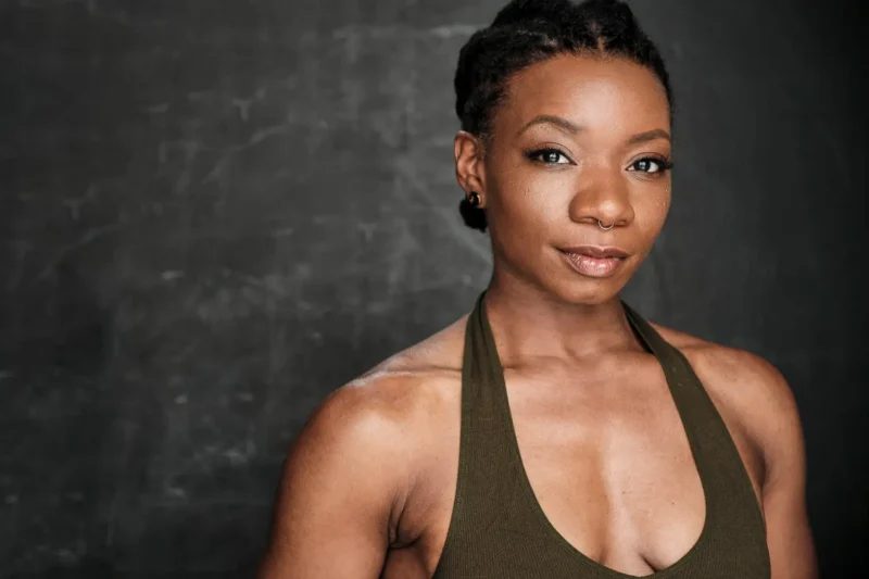 A woman with short braided hair, wearing a sleeveless olive green top, stands in front of a dark, textured background. She is looking slightly to the side with a neutral expression.