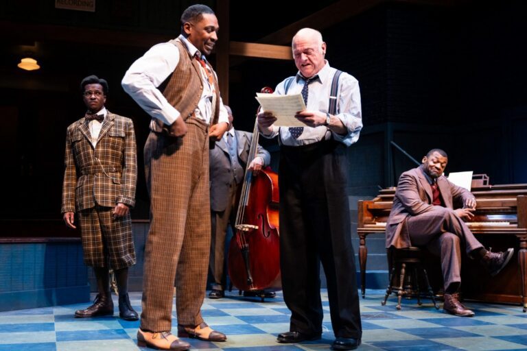 Four men in period clothing stand on a stage, one reading a paper aloud, while a fifth man sits at a piano in the background. A double bass is also visible.