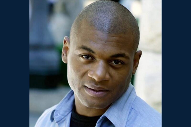A man with short hair and a slight smile looks at the camera, wearing a light blue collared shirt over a dark top. The background is blurred.