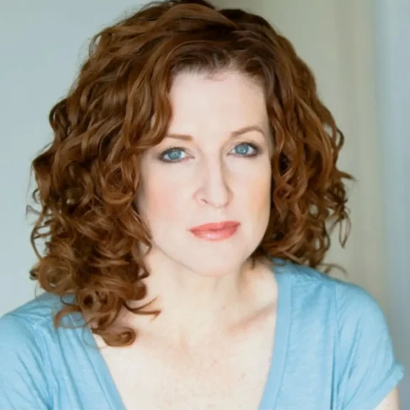 A woman with curly red hair and blue eyes wears a light blue top and looks directly at the camera with a neutral expression.