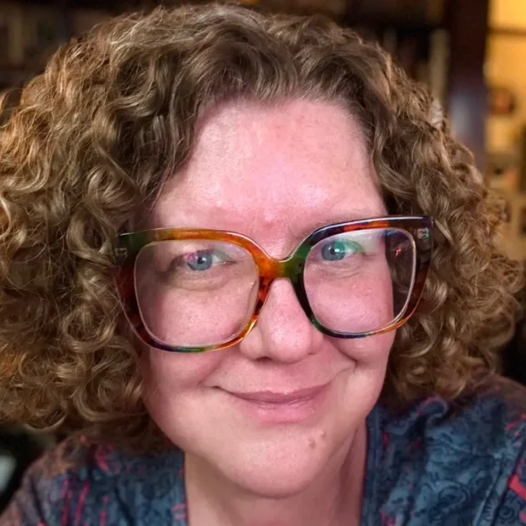 A person with short curly hair and large, multicolored glasses smiles at the camera, wearing a patterned shirt.