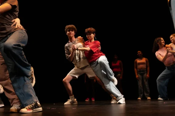 Two people dance together on stage, surrounded by other dancers in casual clothing, with a dark background and stage lighting.
