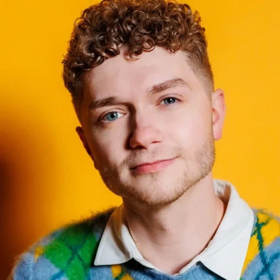 A young person with short curly hair and light eyes is wearing a patterned sweater over a collared shirt, posing in front of a bright yellow background.