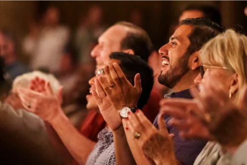 A group of people sit closely together, smiling and clapping, appearing to enjoy a live performance or event.