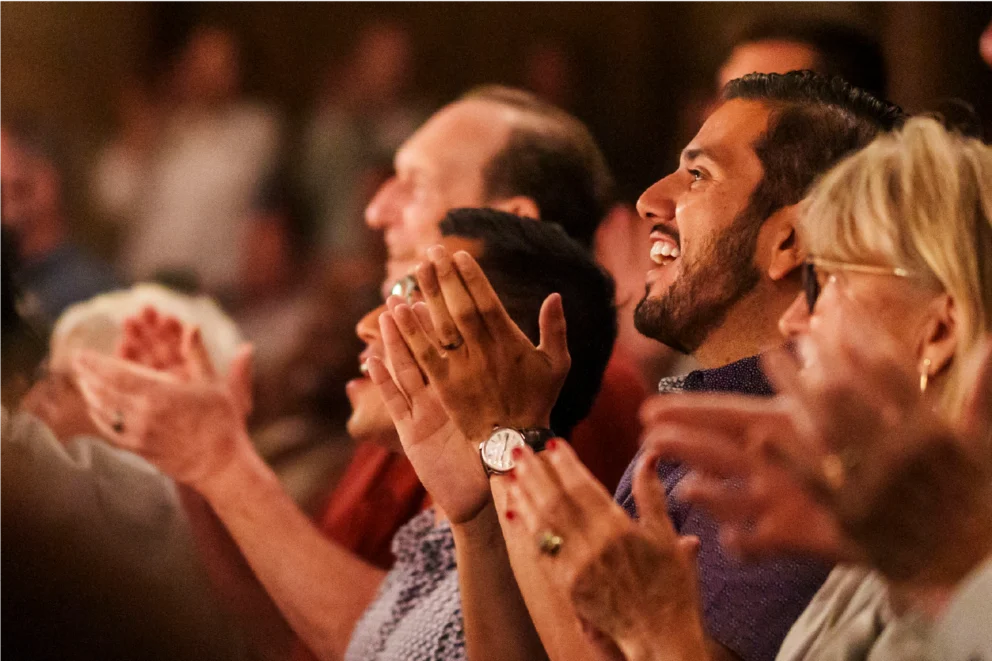 A group of people sit closely together, smiling and clapping, appearing to enjoy a live performance or event.