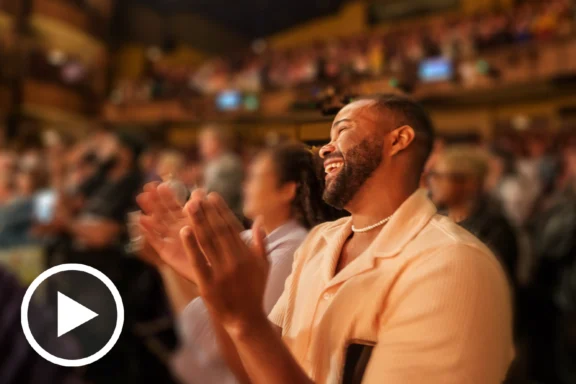A man in a light shirt smiles and applauds in a crowded theater. The audience around him is blurred, and a play button icon is overlaid in the lower left corner.