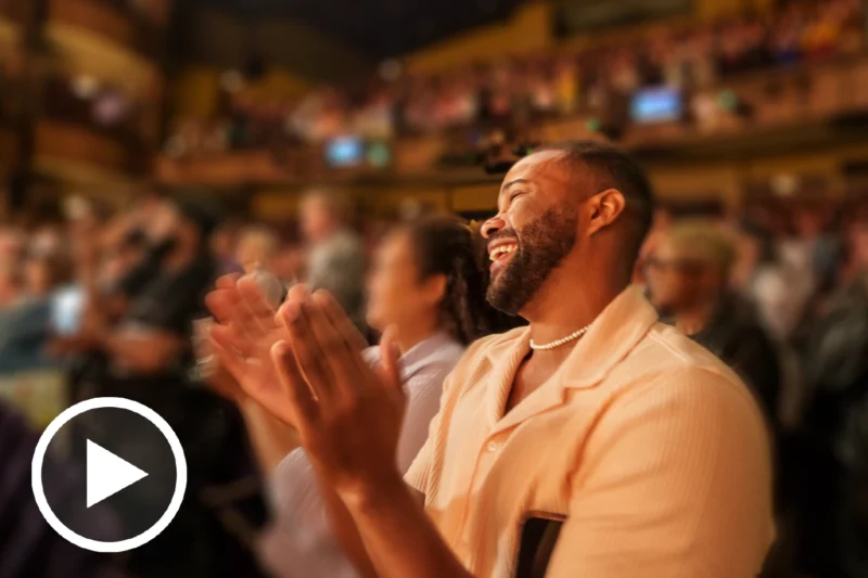 A man in a light shirt smiles and applauds in a crowded theater. The audience around him is blurred, and a play button icon is overlaid in the lower left corner.
