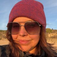 Woman wearing large sunglasses and a purple knit beanie, outdoors with sunlight on her face and a rocky background.