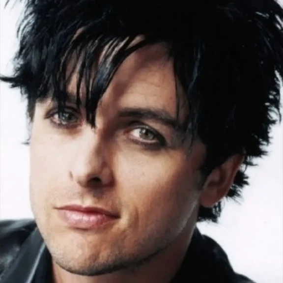 A person with fair skin, green eyes, and black, tousled hair looks at the camera. They are wearing subtle eyeliner and a black jacket.