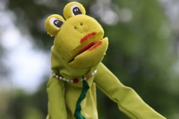A green frog puppet with red lips, a beaded necklace, and a green tie is positioned outdoors with a blurred, leafy background.