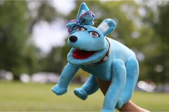 A hand puppet of a blue dog with a pink mouth, white teeth, cartoon eyes, and a sparkly blue bow is held outdoors against a blurred green background.