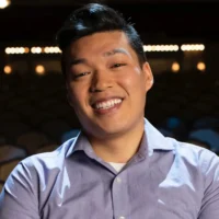 A man in a light purple collared shirt smiles while sitting in an empty theater with rows of seats and stage lights in the background.