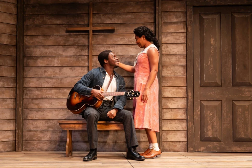A man holding a guitar sits on a bench, looking up and smiling at a woman standing beside him, inside a wooden room with a cross on the wall.