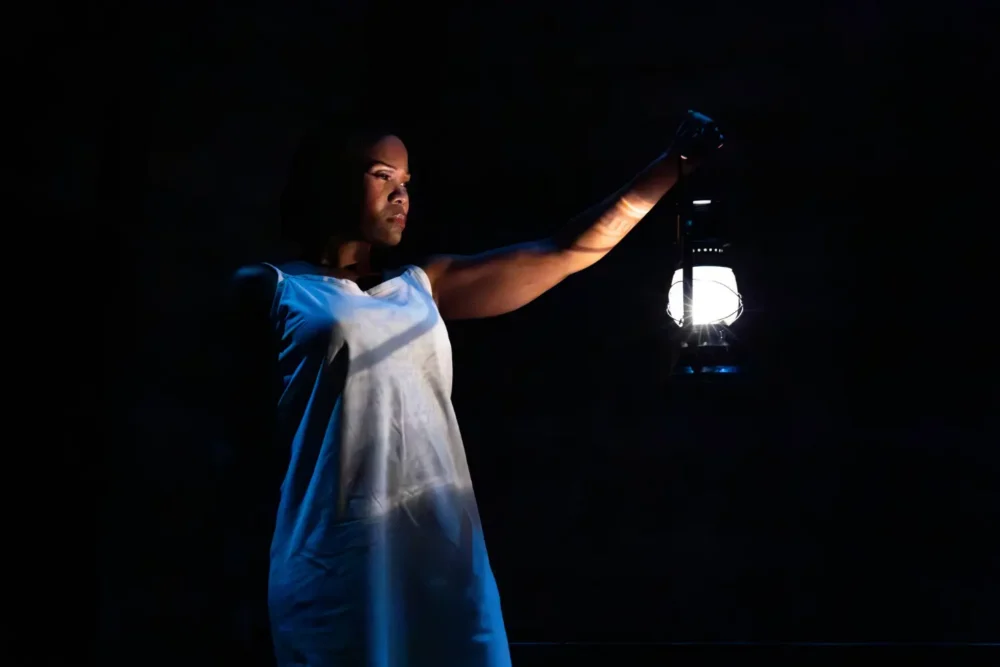 A person in a white dress stands in a dark space, holding a lit lantern aloft, illuminating their face and upper body.